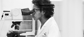 A black and white image of a female scientist in a lab coat and clear protective glasses looking into a microscope
