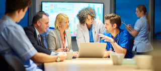 A meeting in a hospital conference room with two healthcare providers in scrubs meeting with other team members in business attire around a laptop with a large screen in the background.
